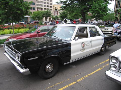 1991 Ford LTD Crown Victoria - black-and-white late-1960s Plymouth police sedan with a roof loudspeaker array and door shield sits at a downtown car show.