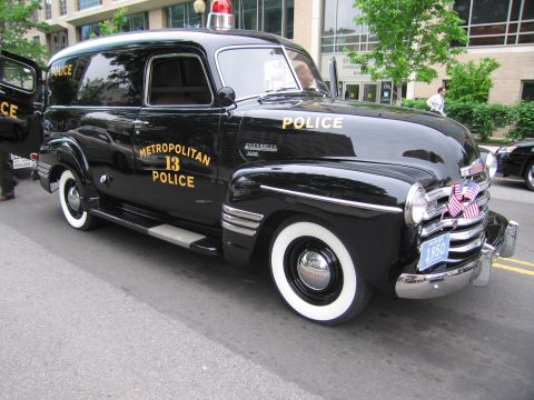 1991 Ford LTD Crown Victoria - restored black 1950 Chevrolet panel delivery truck lettered METROPOLITAN POLICE 13 with whitewall tires sits on a city street.