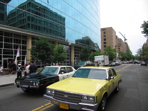 1991 Ford LTD Crown Victoria - yellow STATE POLICE sedan and a black-and-white police cruiser are parked along a city street beside a glass office tower.