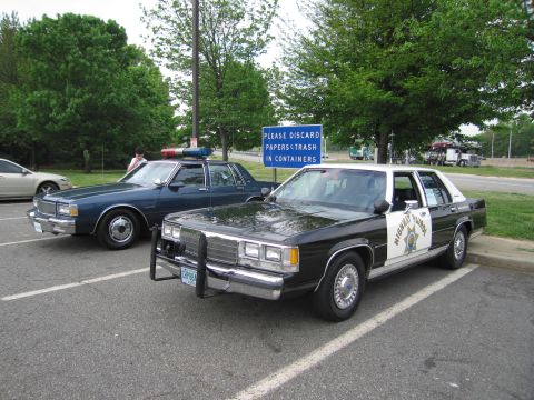 1991 Ford LTD Crown Victoria - black-and-white late-1980s Ford LTD highway patrol sedan and a blue Chevrolet Caprice cruiser sit in a tree-lined parking lot.