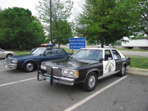 1991 Ford LTD Crown Victoria - black-and-white Ford highway patrol sedan with a push bar and a blue Chevrolet Caprice police car sit in a parking lot.