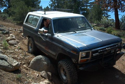 1986 Ford Bronco - A person drives a blue Ford Bronco on a dirt path surrounded by rocks and trees.