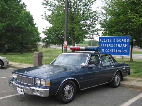 1991 Ford LTD Crown Victoria - dark blue late-1980s Chevrolet Caprice police sedan with a roof lightbar is parked in a lot near a blue trash-disposal sign.