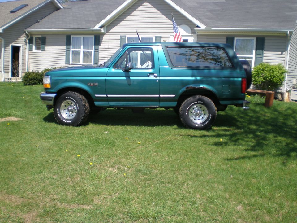 Teal Ford Bronco SUV side view on grass in front of a tan house with American flag