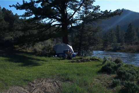 1986 Ford Bronco - A campsite is set up next to a flowing river, surrounded by trees and hills.