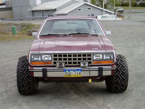 2000 Ford E350 - A maroon AMC Eagle SUV with oversized, aggressive tires is parked on a gravel lot in front of a building. The vehicle has a chrome grille,…