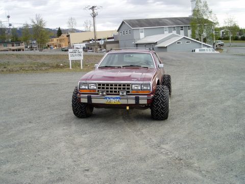 2000 Ford E350 - A lifted, maroon AMC Eagle with oversized tires is parked on a gravel lot in front of a large grey building. A white limousine is parked in…