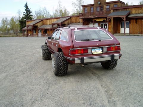2000 Ford E350 - A lifted, maroon AMC Eagle wagon with oversized tires is parked on a gravel lot in front of a wooden building. The rear view shows the vehi…