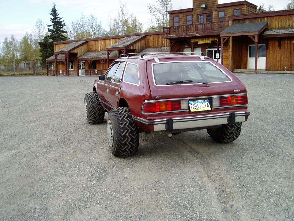 Maroon AMC Eagle wagon with large tires parked in front of a wooden building.