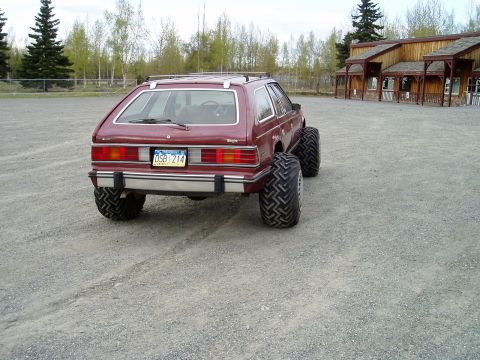 2000 Ford E350 - The rear view of a lifted, maroon AMC Eagle wagon with oversized off-road tires is shown parked on a gravel lot. A wooden building with mul…