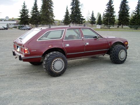 2000 Ford E350 - A maroon station wagon with oversized off-road tires is parked on a gravel lot with trees in the background.
