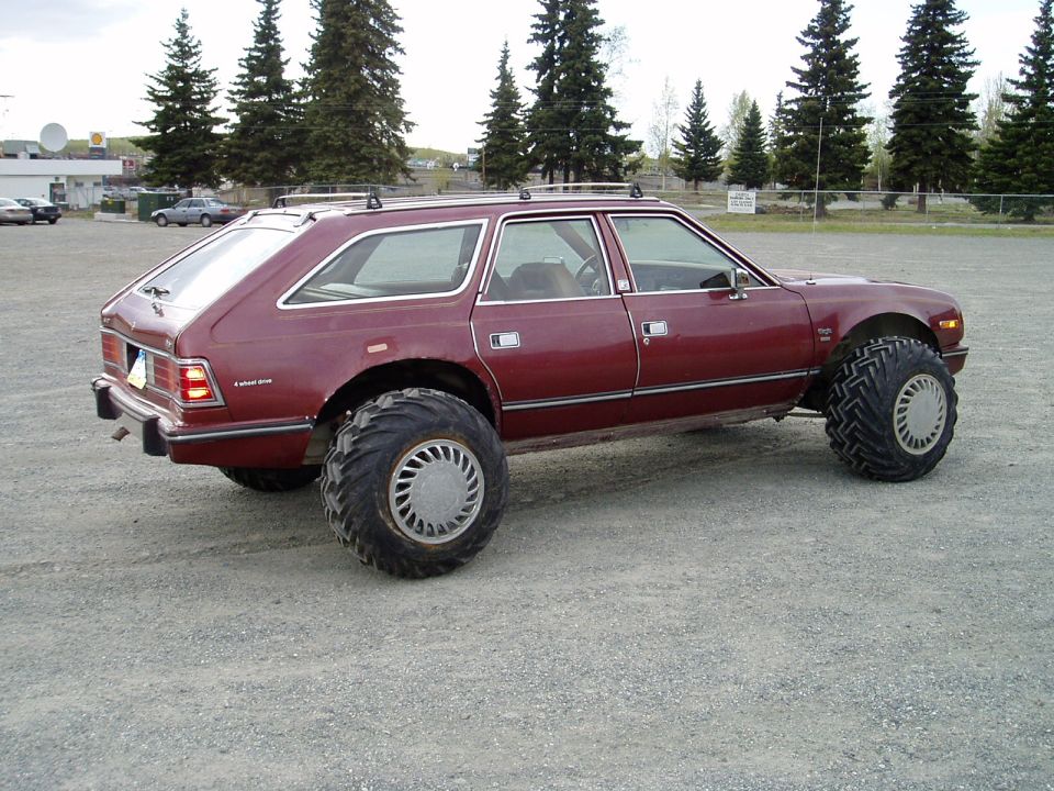 Maroon station wagon with massive tires parked on gravel.