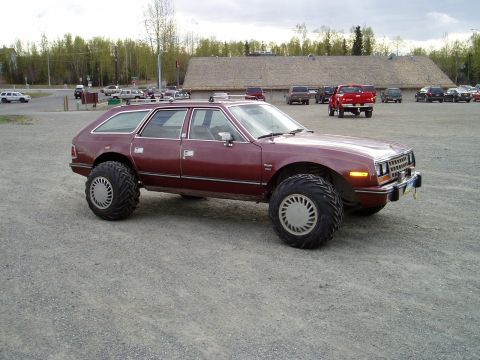 2000 Ford E350 - A lifted, maroon AMC Eagle wagon with oversized tires sits parked on a gravel lot in front of a building.