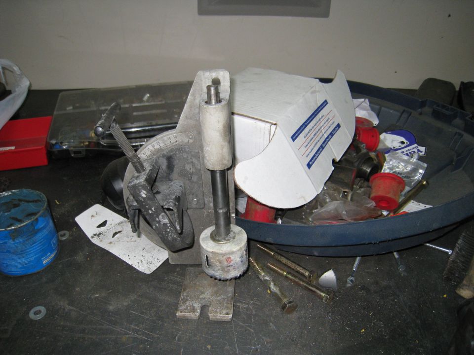 Hole saw jig with a hole saw attached, sitting on a workbench with various parts and tools in a bin