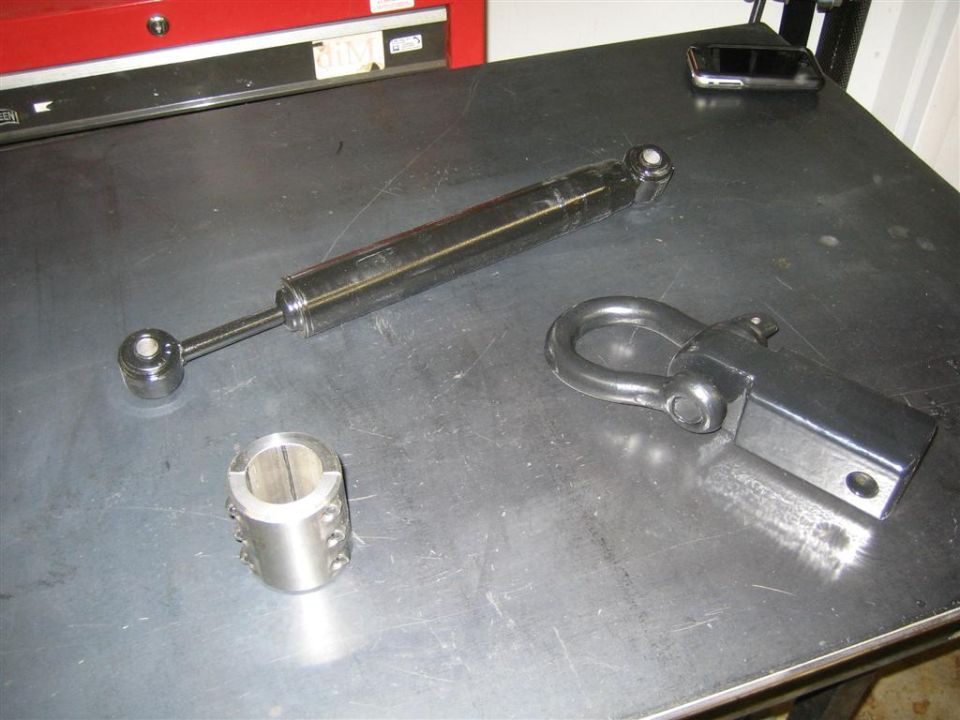 Stainless steel shock absorber, black tow hitch receiver with shackle, and metal collar on a workbench.