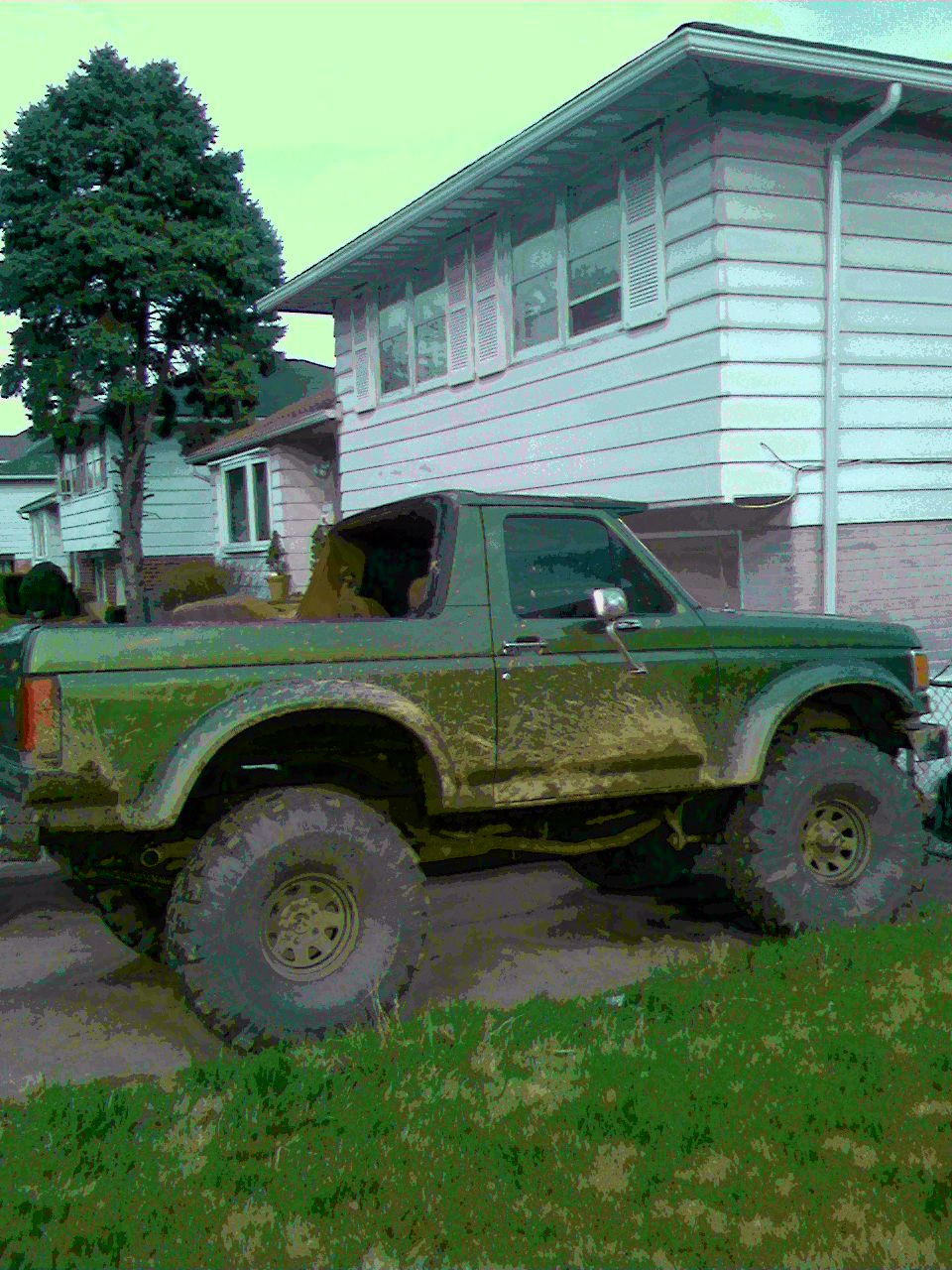 Green lifted Ford Bronco with top off, big mud tires, parked by houses