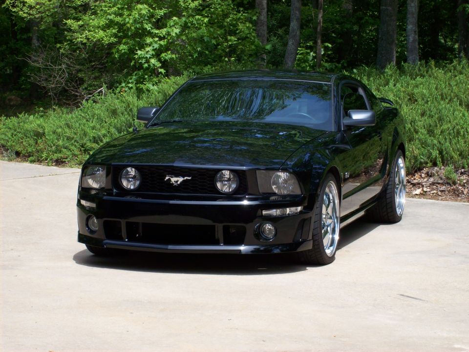 Black Ford Mustang coupe, front 3/4 view, chrome wheels, on driveway near greenery