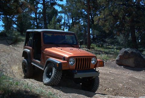 1986 Ford Bronco - An orange Jeep Wrangler is parked on a dirt road surrounded by trees and rocks.