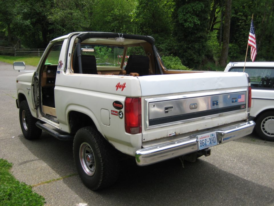 White 1980s Ford Bronco rear passenger side view with American flag on tailgate.