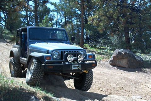1986 Ford Bronco - A blue Jeep Wrangler navigates a dirt path through a wooded area, with a large rock visible to the right.
