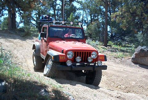 1986 Ford Bronco - A red Jeep Wrangler navigates a dusty trail surrounded by trees and large rocks.