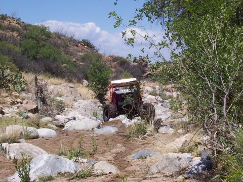 1981 Ford F-150 - A red off-road buggy makes its way through a , rocky terrain with sparse vegetation under a blue sky.