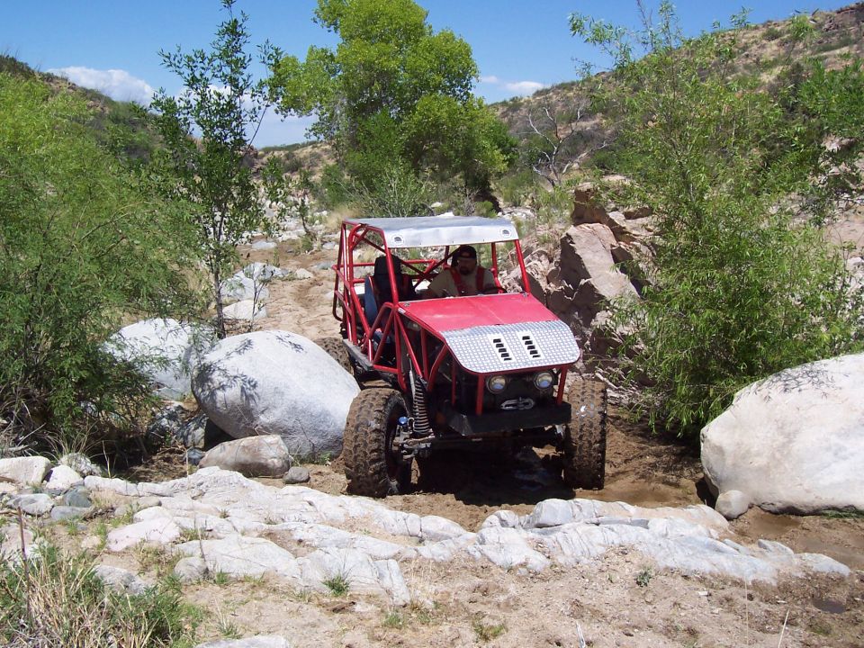Red buggy with roll cage driving over rocks in a dry creek bed. Driver visible