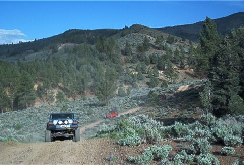 1986 Ford Bronco - Two off-road vehicles navigate a dusty trail through a mountainous landscape under a clear blue sky.