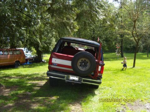 1989 Ford Bronco - A red Ford Bronco is parked in a grassy area with trees and another vehicle in the background.