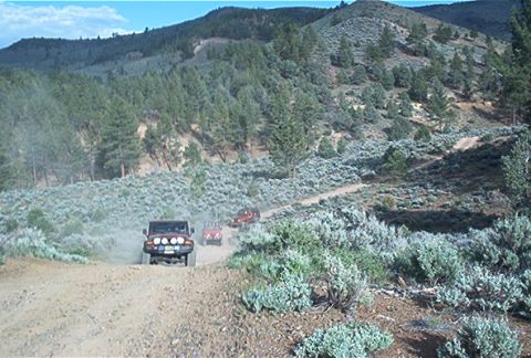 1986 Ford Bronco - Multiple off-road vehicles navigate a dusty trail through a mountainous, arid landscape.