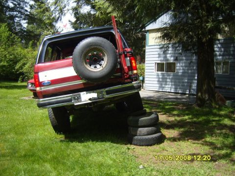 1989 Ford Bronco - Rear view of a red and black Ford Bronco tilted on a stack of tires, with a house and trees in the background.