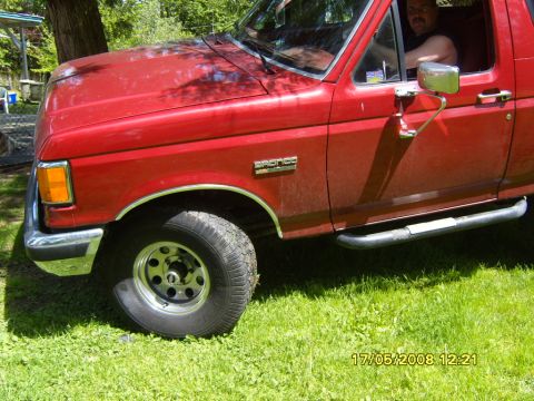 1989 Ford Bronco - A red Ford Bronco is shown from the front quarter angle, parked on a grassy area with trees in the background.