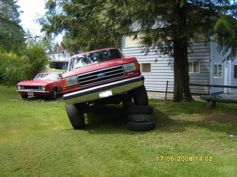 1989 Ford Bronco - Red 1989 Ford Bronco is propped up on tires in a grassy yard next to a classic car.