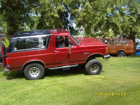 1989 Ford Bronco - Side view of a red 1989 Ford Bronco with a black removable top, parked outdoors with a brown van behind it.
