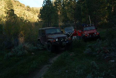 1986 Ford Bronco - Multiple off-road vehicles are parked on a dirt path in a wooded, hilly area.