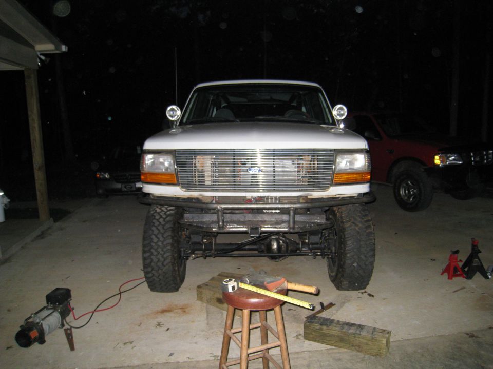 Front view of a white 1996 Ford Bronco with a custom metal bumper and off-road tires. Another red truck is parked behind it
