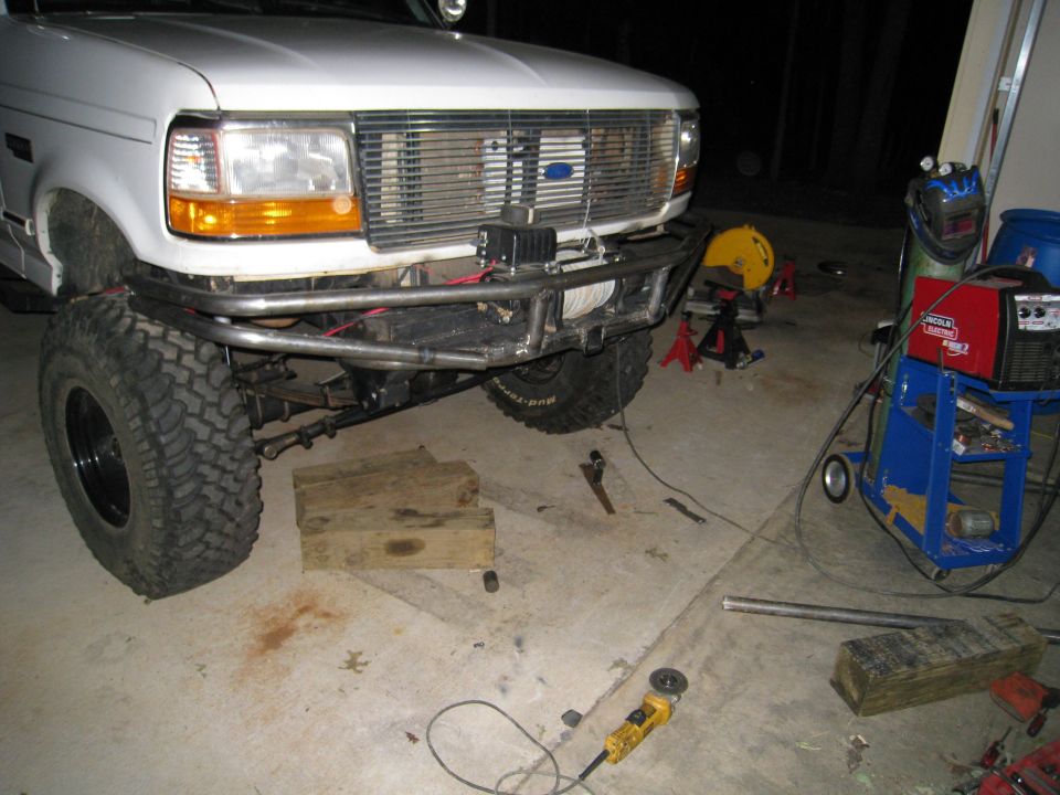 White Ford truck front end with custom metal bumper and winch. Grille has a blue Ford logo. Tools and welding equipment are in the background
