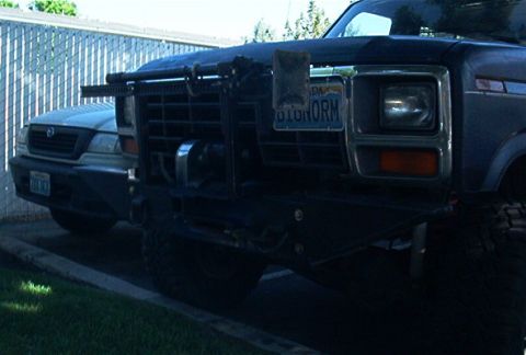 1986 Ford Bronco - A dark Ford truck from the early 1980s is parked next to another vehicle, showing its front grille and winch.