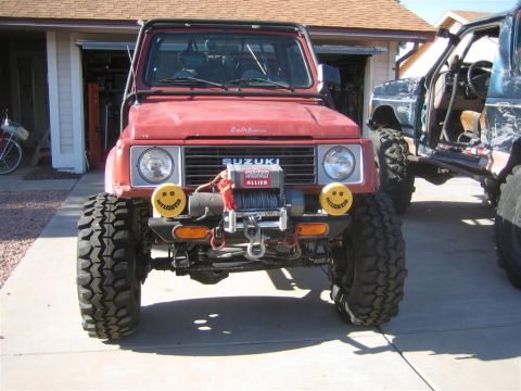 1987 Suzuki Samurai - A red 1987 Suzuki Samurai is parked in front of a garage, next to another off-road vehicle.