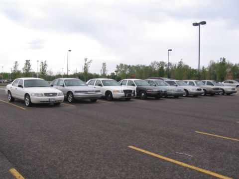 1988 Mercury Grand Marquis - A line of Ford Crown Victoria police cars are parked in a lot under an overcast sky.