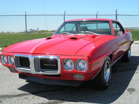 1969 Pontiac Firebird - A red 1969 Pontiac Firebird is parked on asphalt with a chain-link fence and a small airplane in the distance.
