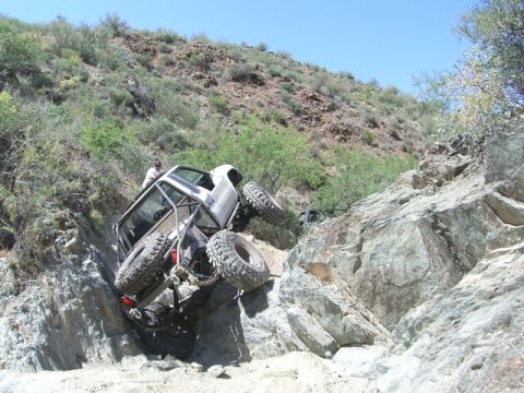 1986 Toyota Hilux - A white Toyota Hilux is shown at a steep angle, navigating a rocky off-road trail. The vehicle has a roll cage and oversized tires, indicat…