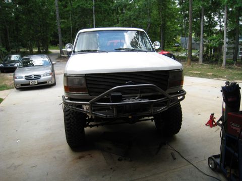 1996 Ford Bronco - A white Ford Bronco with a custom metal bumper and winch is parked on a concrete driveway in front of a wooded area. Two sedans are parked…