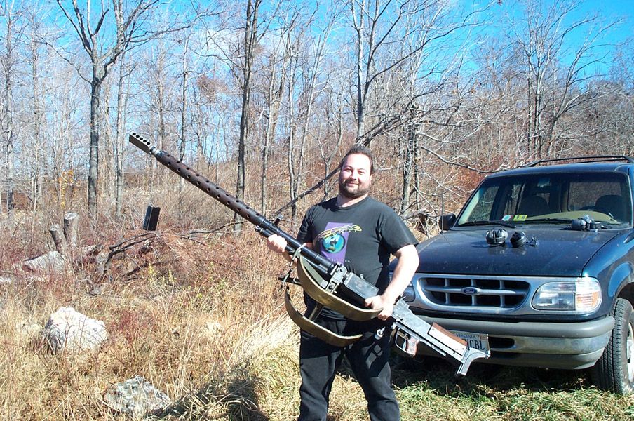 Man holding a large machine gun in front of a dark blue Ford Explorer SUV.