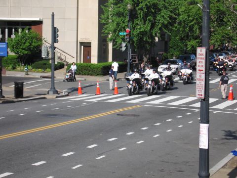1991 Ford LTD Crown Victoria - Police motorcycle officers ride toward a coned-off crosswalk on a wide city street lined with parked cars and signage.