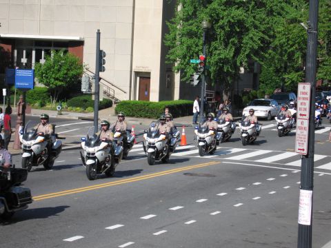 1991 Ford LTD Crown Victoria - group of police officers in tan uniforms ride motorcycles through a downtown intersection with traffic cones and pedestrians.