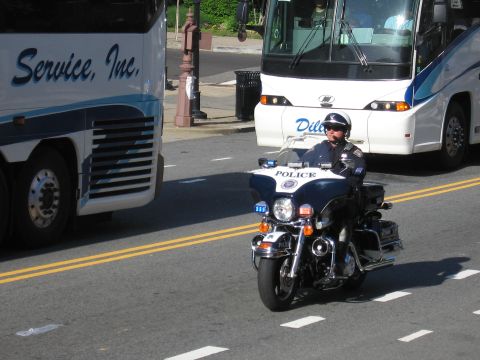 1991 Ford LTD Crown Victoria - helmeted officer rides a black-and-white Harley-Davidson marked POLICE down a street alongside parked tour buses.