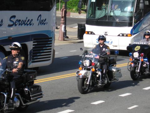 1991 Ford LTD Crown Victoria - Several police motorcycle officers ride together down a city street past tour buses and a brick building.