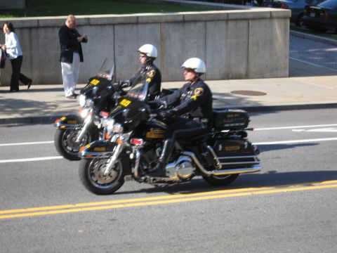1991 Ford LTD Crown Victoria - Two police motorcycle officers in white helmets ride black Harley-Davidsons along a street beside a stone retaining wall.