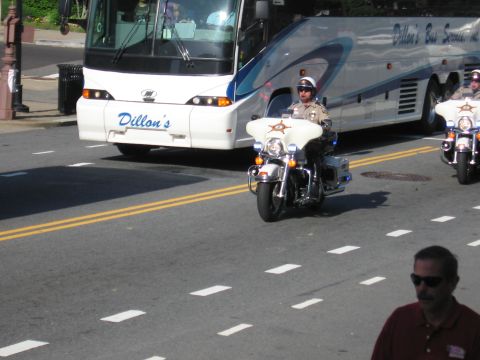 1991 Ford LTD Crown Victoria - sheriff's motorcycle officer rides beside a white Dillon's motorcoach as a man in sunglasses watches in the foreground.